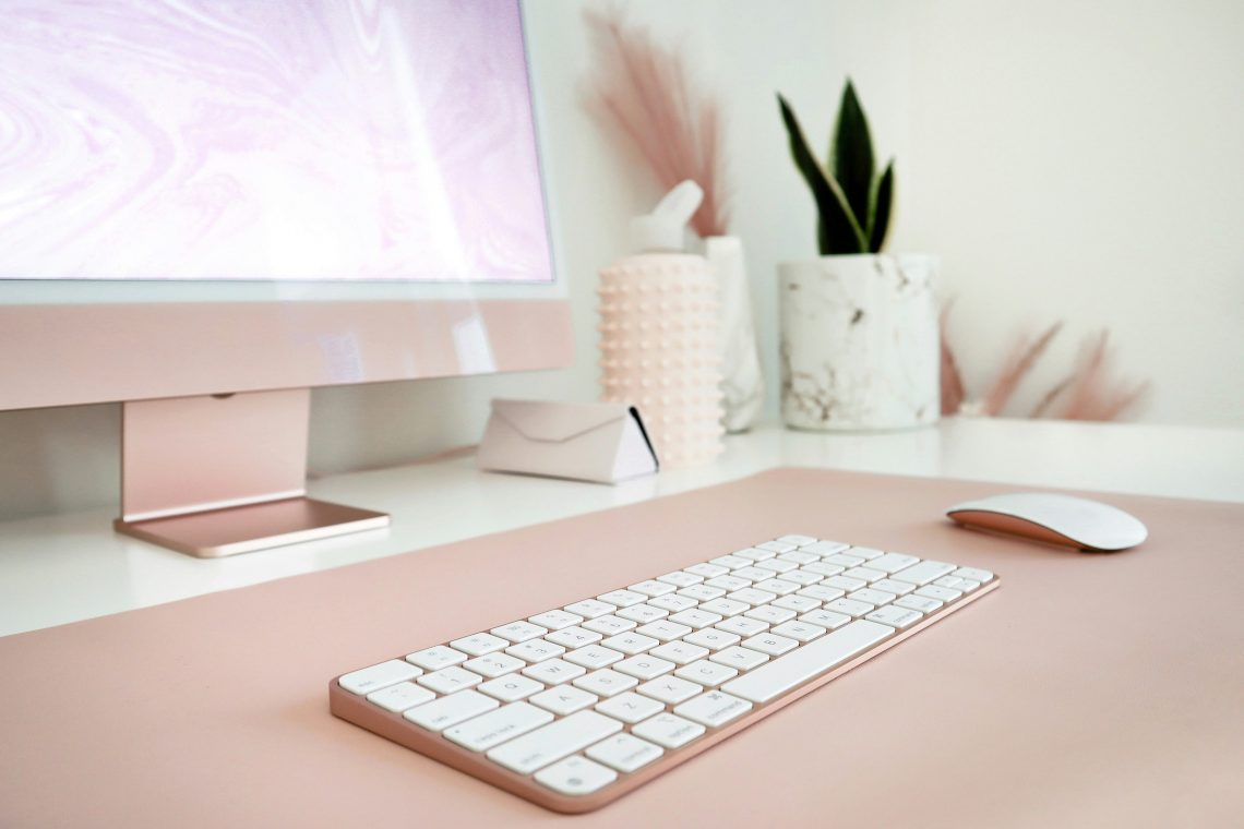 Low angle shot of desk with pink pc screen, keyboard and mouse. glasses case, plant and light desk décor in back right of desk