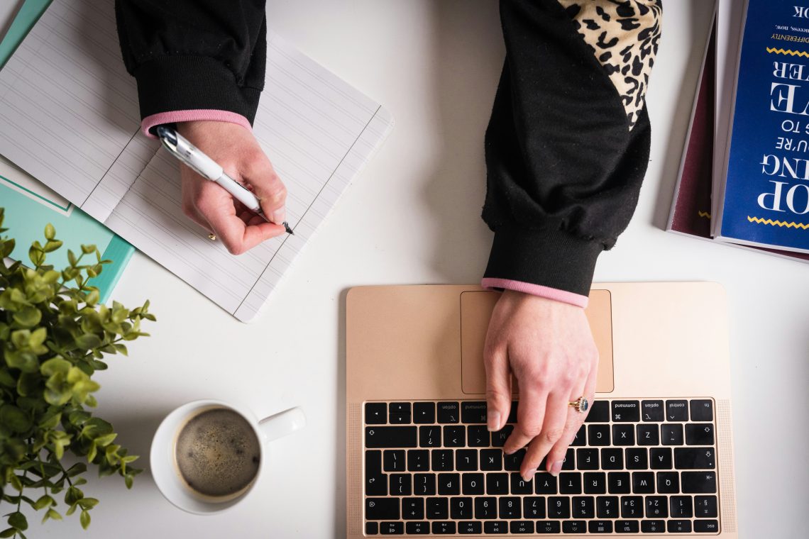 overhead shot of desk, female hands on laptop keyboard and pen in other hand about to write.